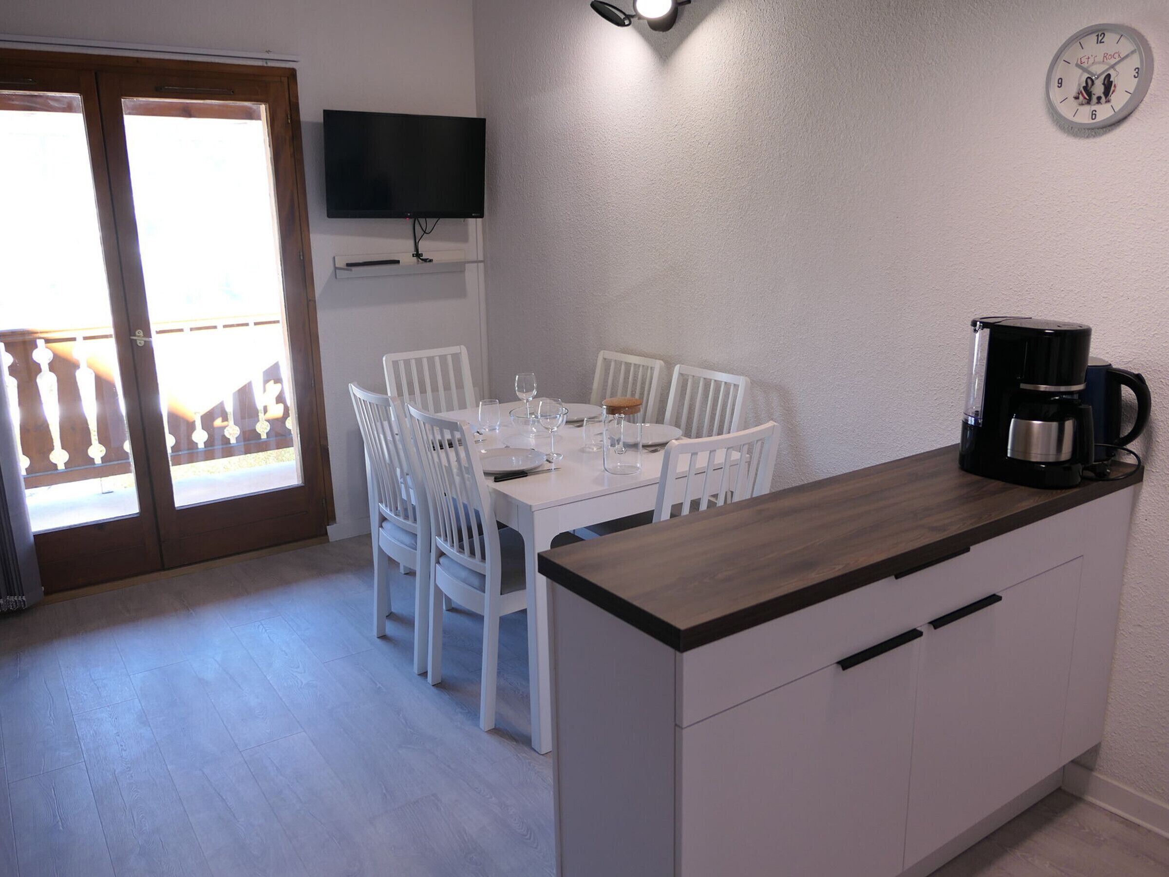 Un coin repas avec une table et des chaises blanches, une machine à café posée sur le comptoir et une horloge au mur.