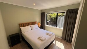 A neatly made bed with white linens and two folded towels, a bedside lamp, and a window with curtains. - Golden Bay Heights - Luxury Accommodation (Parapara)