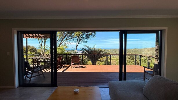 A room with a sliding glass door leading to an outdoor deck with a wooden table and chairs, overlooking a scenic landscape. - Golden Bay Heights - Luxury Accommodation (Parapara)