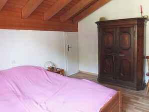 A bedroom with a wooden ceiling, a bed with a pink bedspread, a wooden wardrobe, and a nightstand with a lamp.