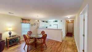 A dining area with a glass table and wicker chairs, a kitchen with white cabinets, and a hallway with a wooden floor. - Pawleys Island Fairway Retreat 3BR Villa (Pawleys Island)