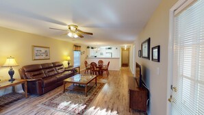 A living room with a brown leather sofa, a glass coffee table, and a dining area with wooden chairs. A ceiling fan is visible, and the room has wooden flooring. - Pawleys Island Fairway Retreat 3BR Villa (Pawleys Island)