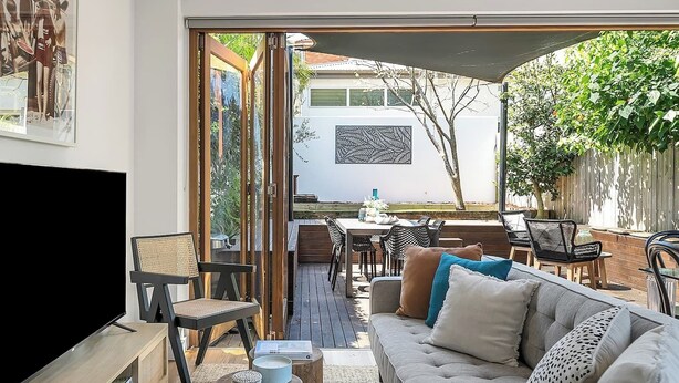 A modern living room with a TV, sofa, and coffee table. There is a dining area with chairs and a table. The room has a large window and a view of the outdoors.