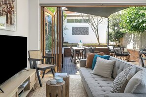 A modern living room with a TV, sofa, and coffee table. There is a dining area with chairs and a table. The room has a large window and a view of the outdoors. - Artscape - Bondi Beach Villa (Bondi Beach)
