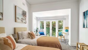 A hotel room with two beds, a sitting area with a chair and table, and a balcony with a view of greenery.