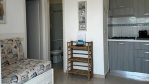 A compact living space with a kitchenette, a patterned sofa, and a wooden shelf unit.