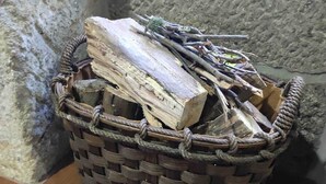Una cesta tejida llena de leña y ramas, sobre un suelo de baldosas frente a una pared de piedra.