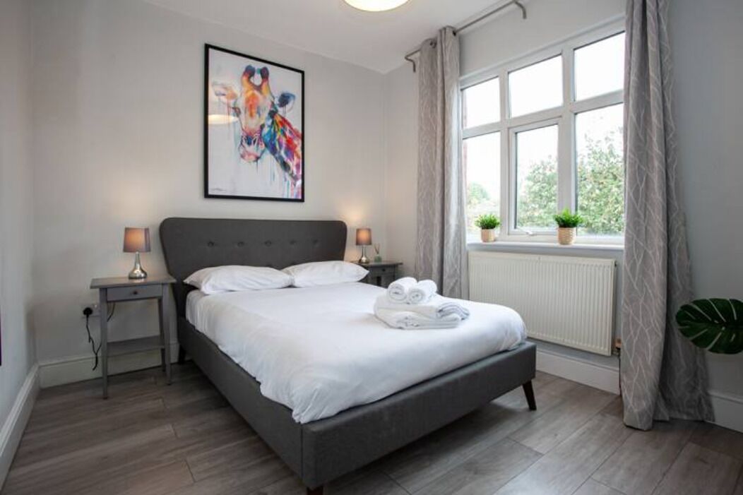 A modern bedroom with a grey bed, white linens, a framed abstract painting, a bedside lamp, a window with curtains, and a potted plant.