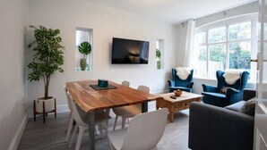 Una sala moderna con una mesa de comedor de madera, sillas blancas, un sillón azul y un sofá gris. Hay un televisor montado en la pared y un ventanal con cortinas blancas.