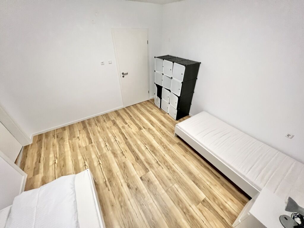 Ett litet, rent rum med trägolv, en vit dörr och en vit säng.