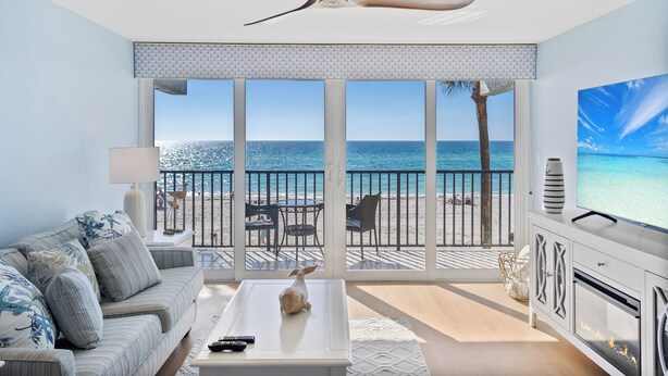 A living room with a large window overlooking a beach, a ceiling fan, and a fireplace. - Luxury Gulf-Front Condo on Longboat Key with Breathtaking Sunsets (Longboat Key)