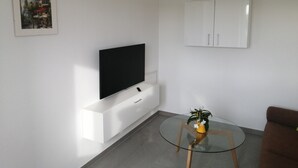 A modern living room with a flat-screen TV mounted on a white cabinet, a glass coffee table with a plant, and a brown sofa.