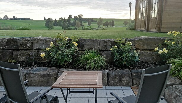 A patio with two chairs and a table, overlooking a stone wall with plants and a view of a house and fields.