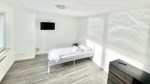 Une petite pièce lumineuse, avec un lit simple, un téléviseur à écran plat fixé au mur et une commode en bois.