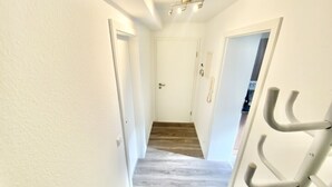 Un couloir étroit aux murs blancs et au plancher en bois, qui mène à une porte au fond.