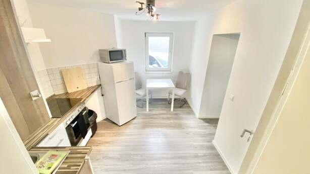 Une cuisine compacte équipée d’un four à micro-ondes, d’un petit réfrigérateur et d’un plan de travail en bois. Une fenêtre se trouve au-dessus d’une table blanche, accompagnée d’une chaise.