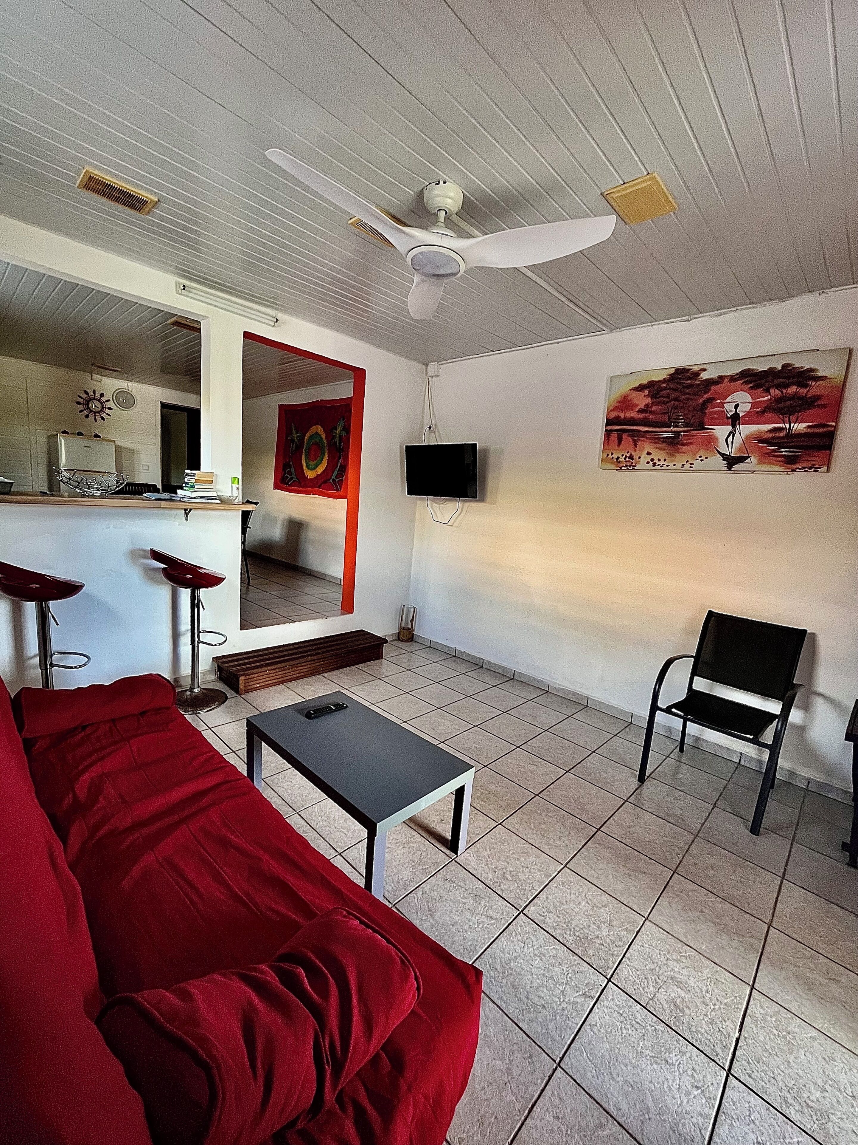 Un salon avec un canapé rouge, un fauteuil noir, une petite table, un ventilateur de plafond, un téléviseur fixé au mur et un tableau.