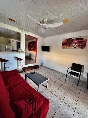 Un salon avec un canapé rouge, un fauteuil noir, une petite table, un ventilateur de plafond, un téléviseur fixé au mur et un tableau.