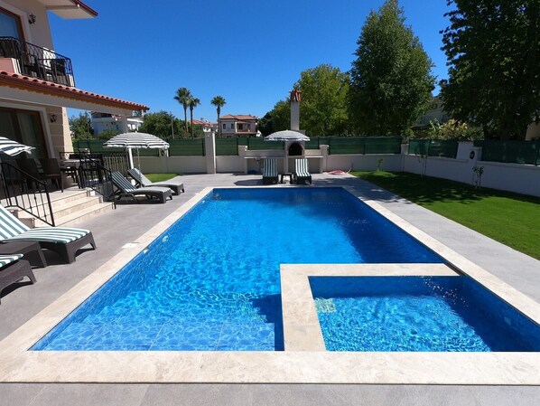 A rectangular swimming pool with a tiled edge, surrounded by a paved deck. There are lounge chairs and umbrellas on the deck.