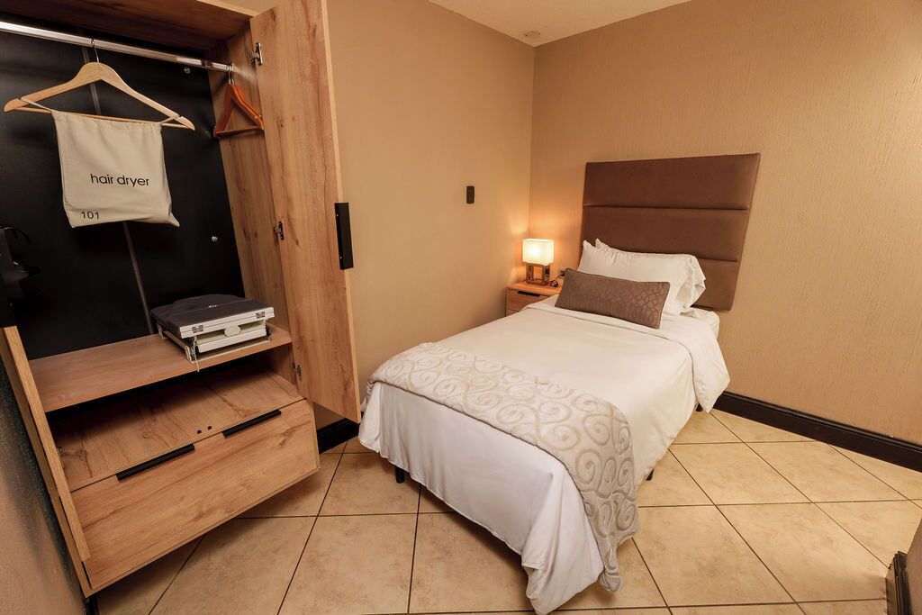 Una habitación de hotel con una cama, un armario de madera con un bolso colgado que dice 'secador de pelo' y una mesita de noche con una lámpara.