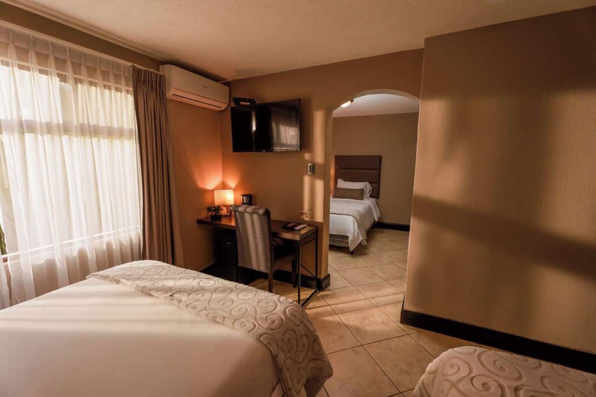 Quarto de hotel com cama, escrivaninha, cadeira, televisão e outra cama ao fundo.