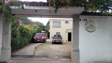Una entrada protegida por una reja con dos coches estacionados en su interior, un edificio con una ventana y una puerta, y un emblema circular en la pared.