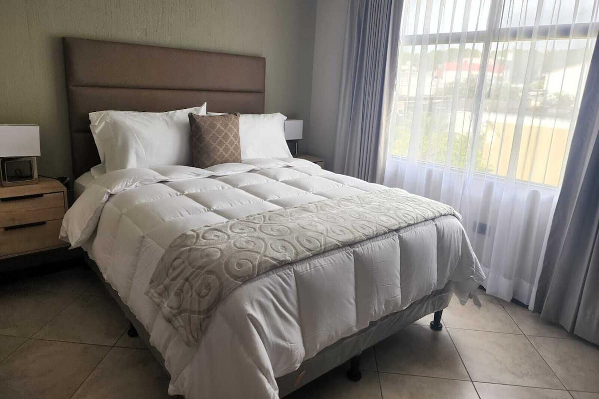 Uma cama bem arrumada com cabeceira, mesa de cabeceira com abajur e janela com cortinas.
