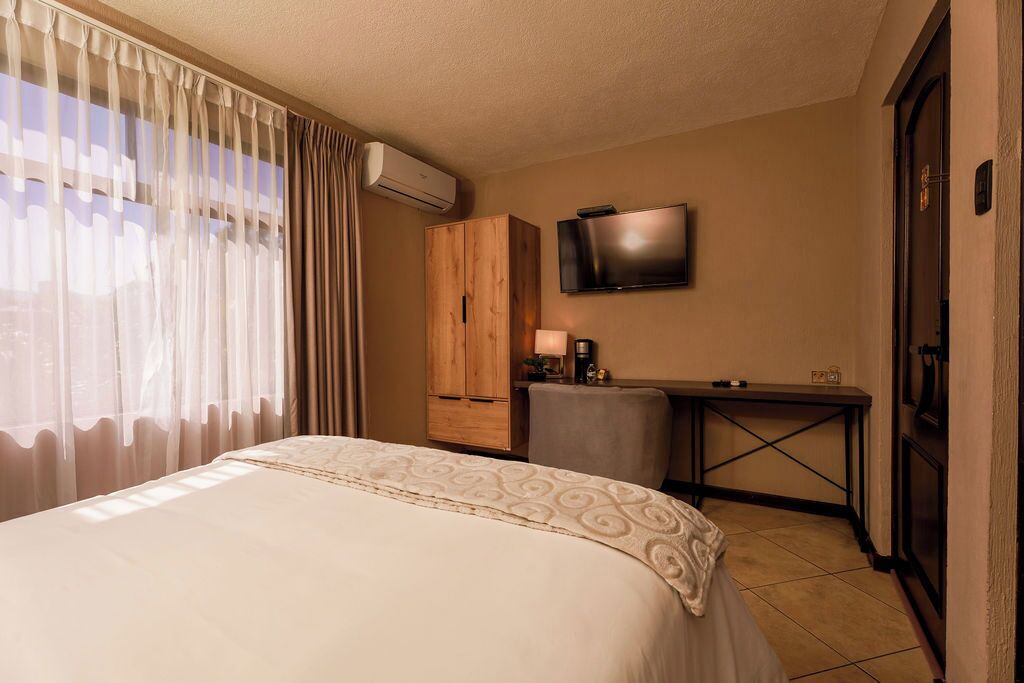A hotel room with a bed, a TV mounted on the wall, a wooden wardrobe, and a small desk with a lamp.