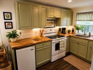 A kitchen with green cabinets, a wooden countertop, a white refrigerator, and a window with blinds.