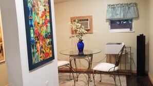 Ein Essbereich mit einem Glastisch, zwei Stühlen, einer blauen Vase mit Blumen, einem kleinen Fenster mit gestreiftem Vorhang und einem Deckenventilator.