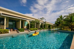 A swimming pool with a person in a yellow kayak, surrounded by palm trees and a building with large windows.