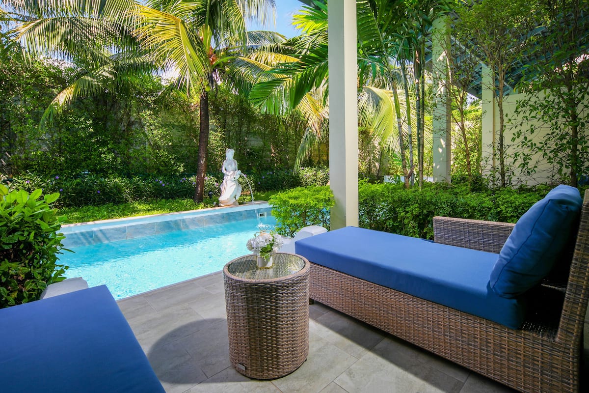 A poolside area with a wicker sofa and a round table, surrounded by lush greenery and a statue.