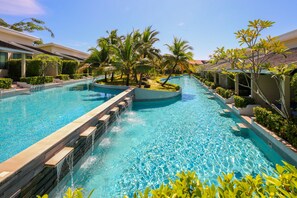 A swimming pool with a waterfall feature, surrounded by palm trees and greenery, with buildings on either side. - CRAFT Resort & Villas (Ratsada)