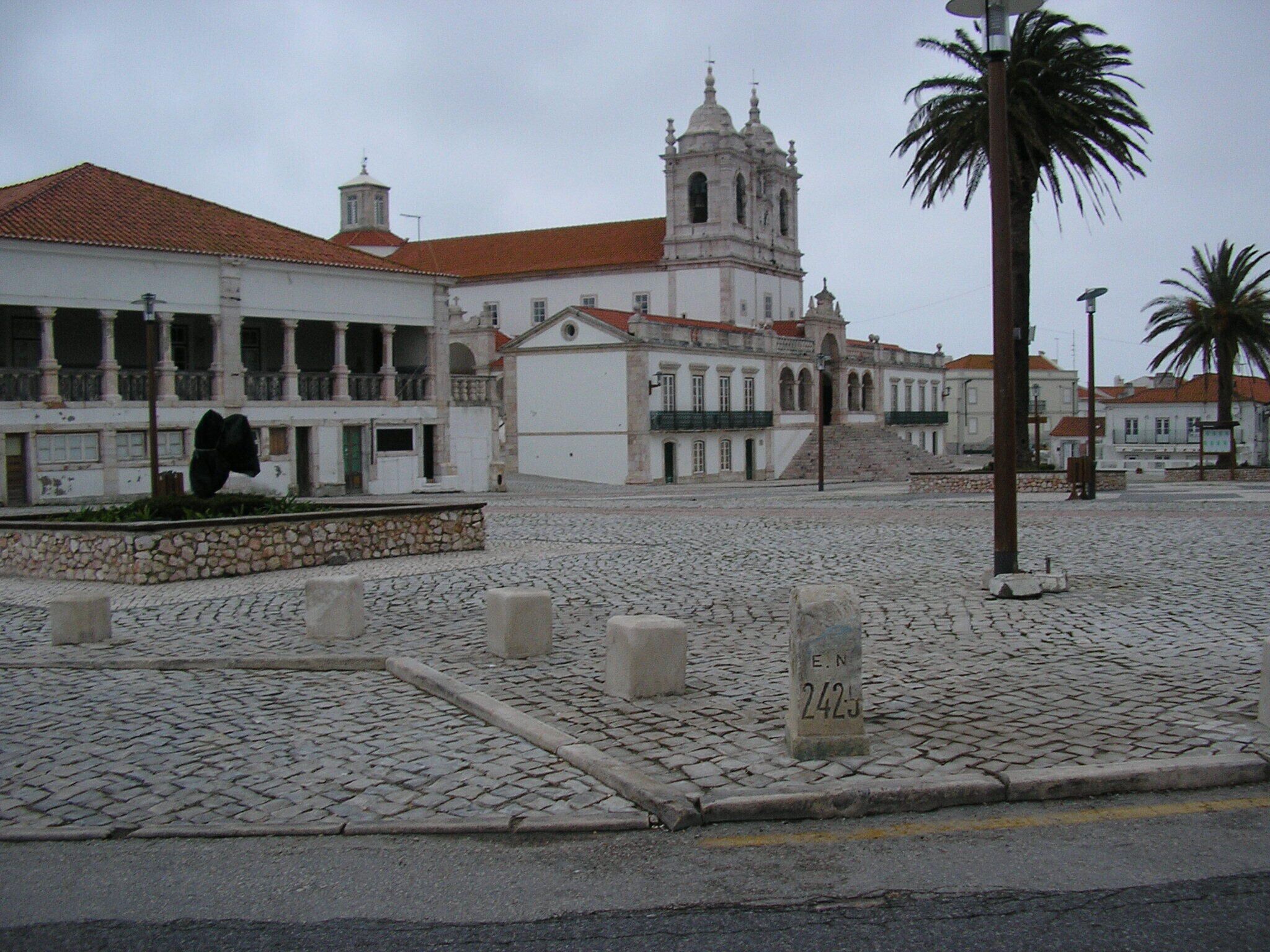 A cobblestone square with historic buildings, including a church with a bell tower, and a palm tree.