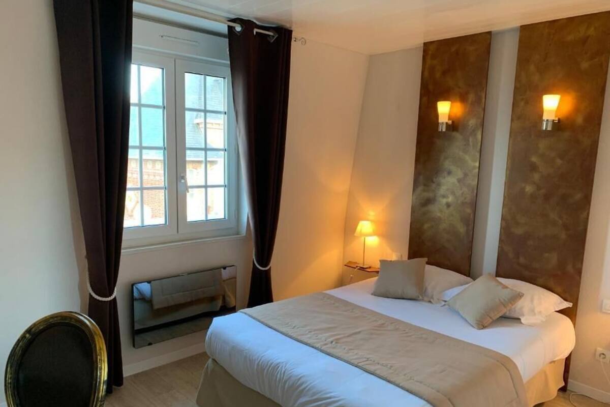A hotel room with a large bed, a bedside table with a lamp, a window with curtains, and a view of the outside.