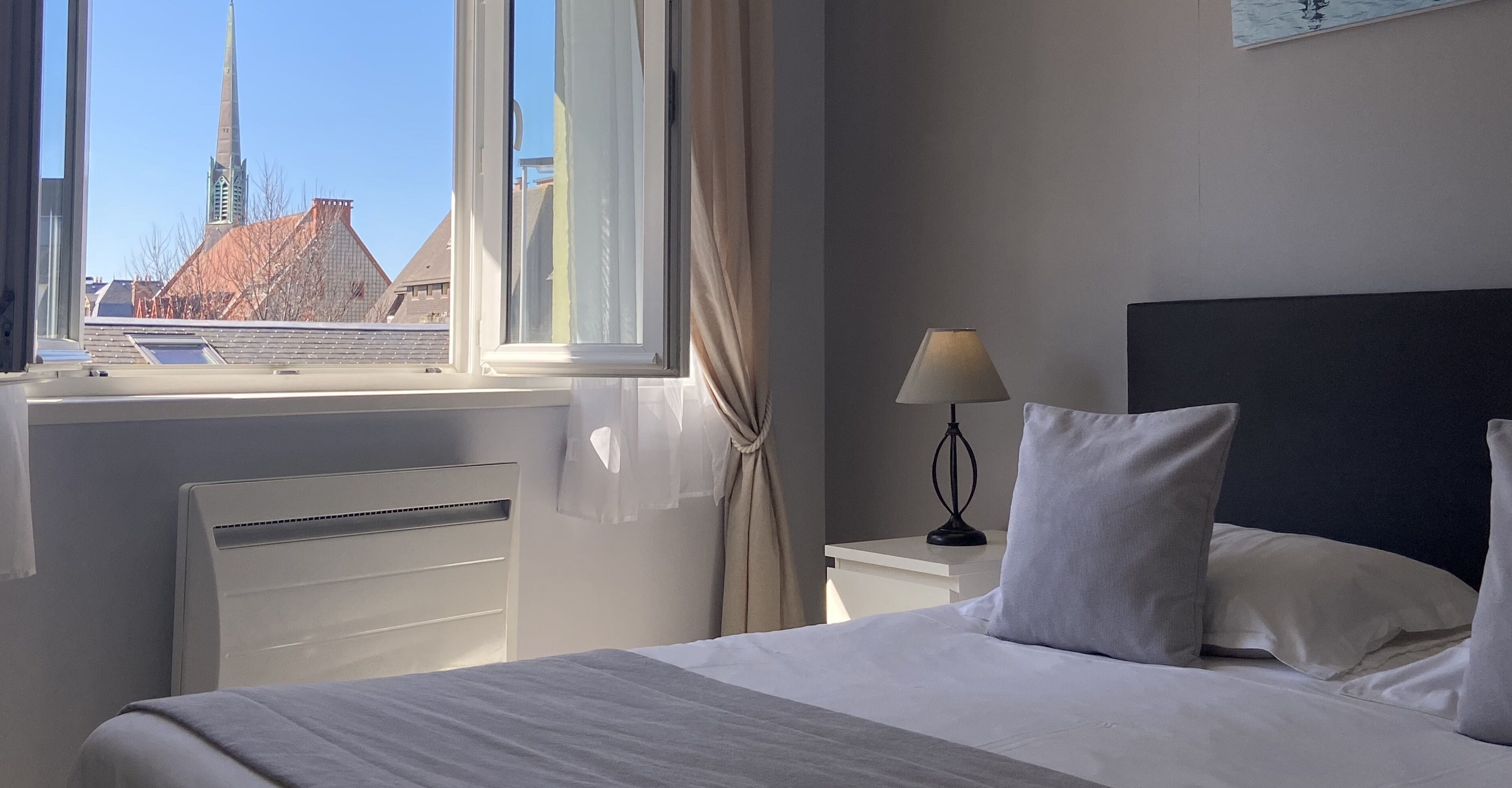 A hotel room with a bed, a bedside table with a lamp, a window with curtains, and a view of a building with a spire.