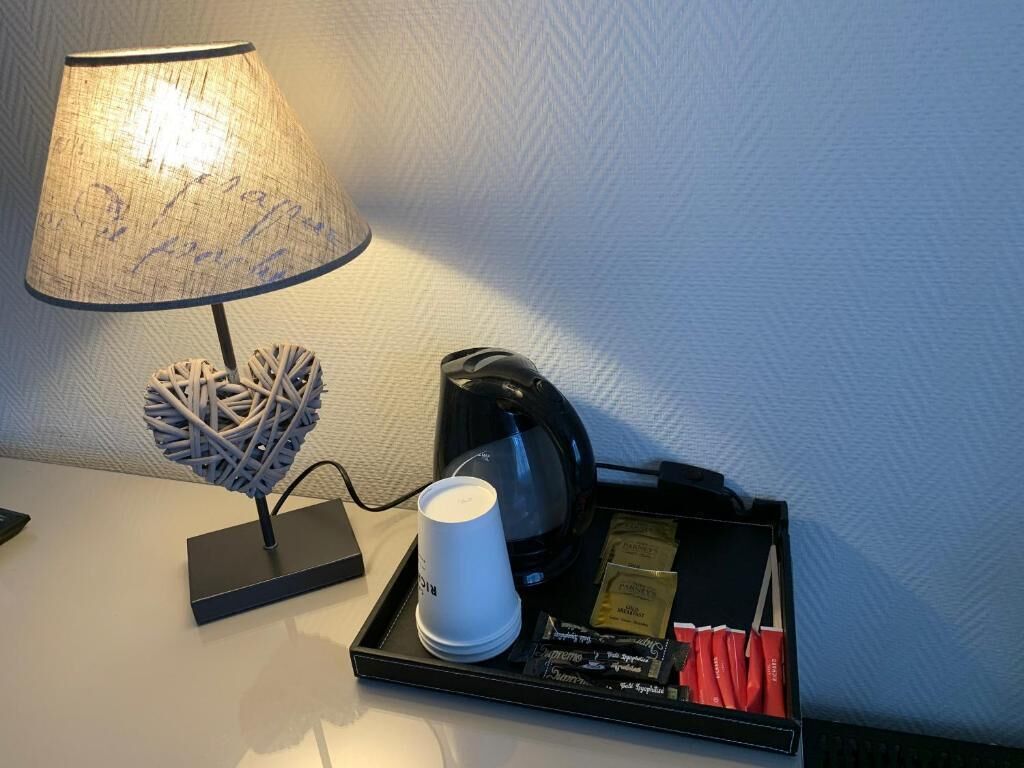 Uma mesa de cabeceira com uma luminária, uma xícara, um tabuleiro com sachês de café e chá, e um objeto decorativo.