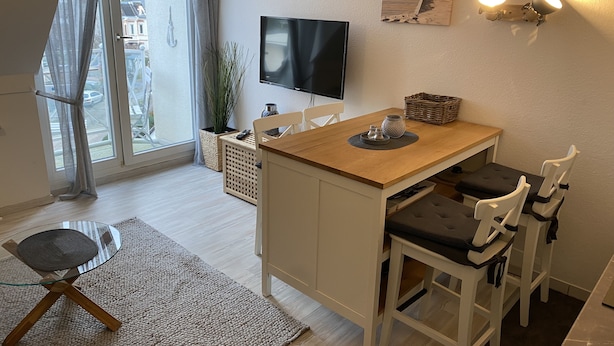 Ein modernes Wohnzimmer mit einem Holzespeisetisch, weißen Stühlen, einem Fernseher und einem großen Fenster mit Vorhängen.