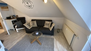 Ein gemütliches Schlafzimmer im Loft-Stil mit einer dunkelgrauen Couch, einem kleinen runden Couchtisch und einem grauen Teppich. Rechts ist eine weiße Heizung sichtbar, und es gibt eine Deckenleuchte.