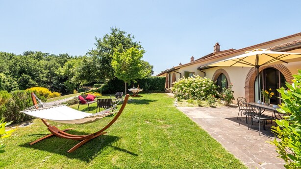 A garden with a hammock, a table with chairs, and a building with a tiled roof.