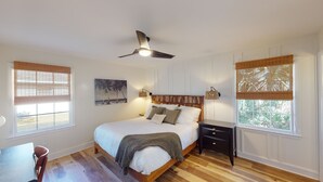 Een slaapkamer met een houten bed, een plafondventilator, twee nachtkastjes en een raam met uitzicht op bomen.