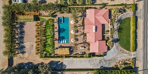 Vista aerea di un resort con piscina, palme e un edificio con tetto rosso.