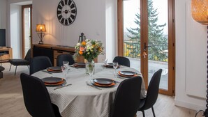 Une salle à manger avec une table ronde dressée pour six personnes, agrémentée de chaises noires, d’une nappe blanche et d’assiettes orange. On peut également voir une grande horloge murale et un vase rempli de fleurs.