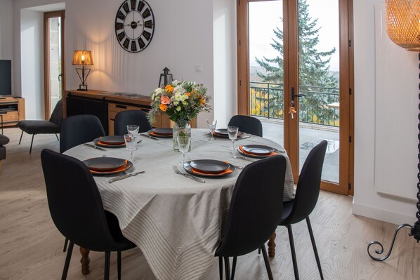 A dining room with a round table set for six, featuring black chairs, white tablecloth, and orange plates. A large wall clock and a vase with flowers are visible.