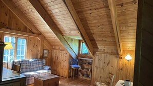 Ein gemütliches Blockhaus mit Schlafzimmer im Obergeschoss, Holzdielen, einer Couch, einem Esstisch und einer kleinen Küchenzeile.
