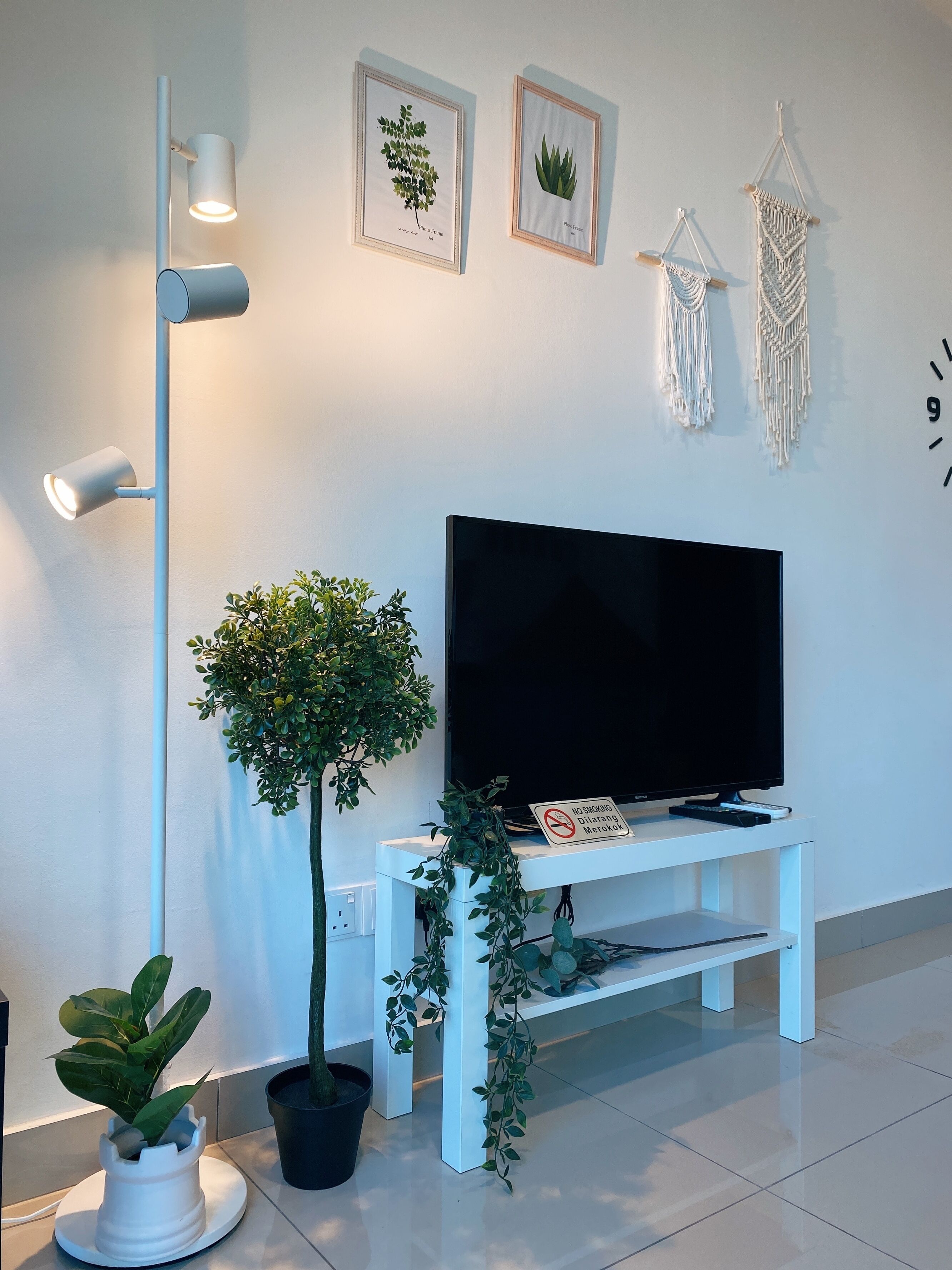 A modern living room with a flat-screen TV on a white stand, a floor lamp, and wall-mounted macrame decorations.