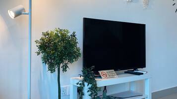 A modern living room with a flat-screen TV on a white stand, a floor lamp, and wall-mounted macrame decorations.