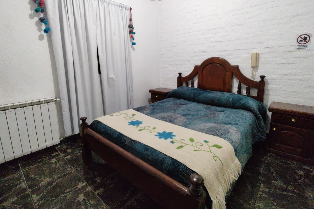 Un dormitorio con una cama de madera, cortinas blancas, un radiador, una mesita de noche y un teléfono instalado en la pared.