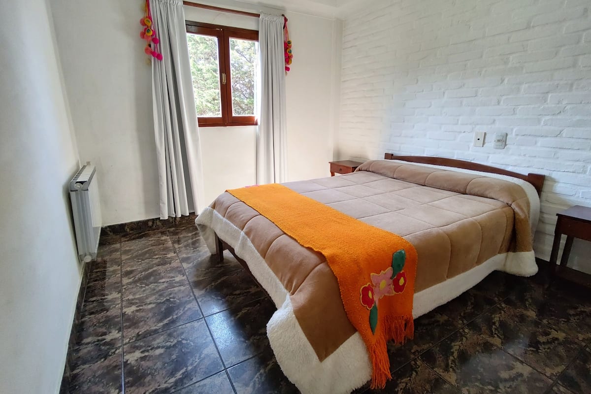 A bedroom with a bed, a window with curtains, a radiator, and a white brick wall.