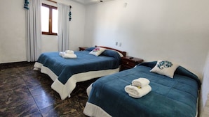 Habitación de hotel con dos camas, cada una con cubrecama azul y almohadas blancas, y una ventana con cortinas.